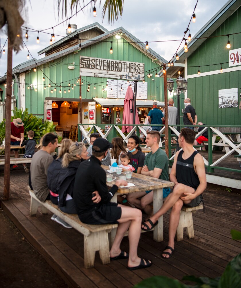A family sits outside on a wooden table at the green Seven Brothers building in Kahuku