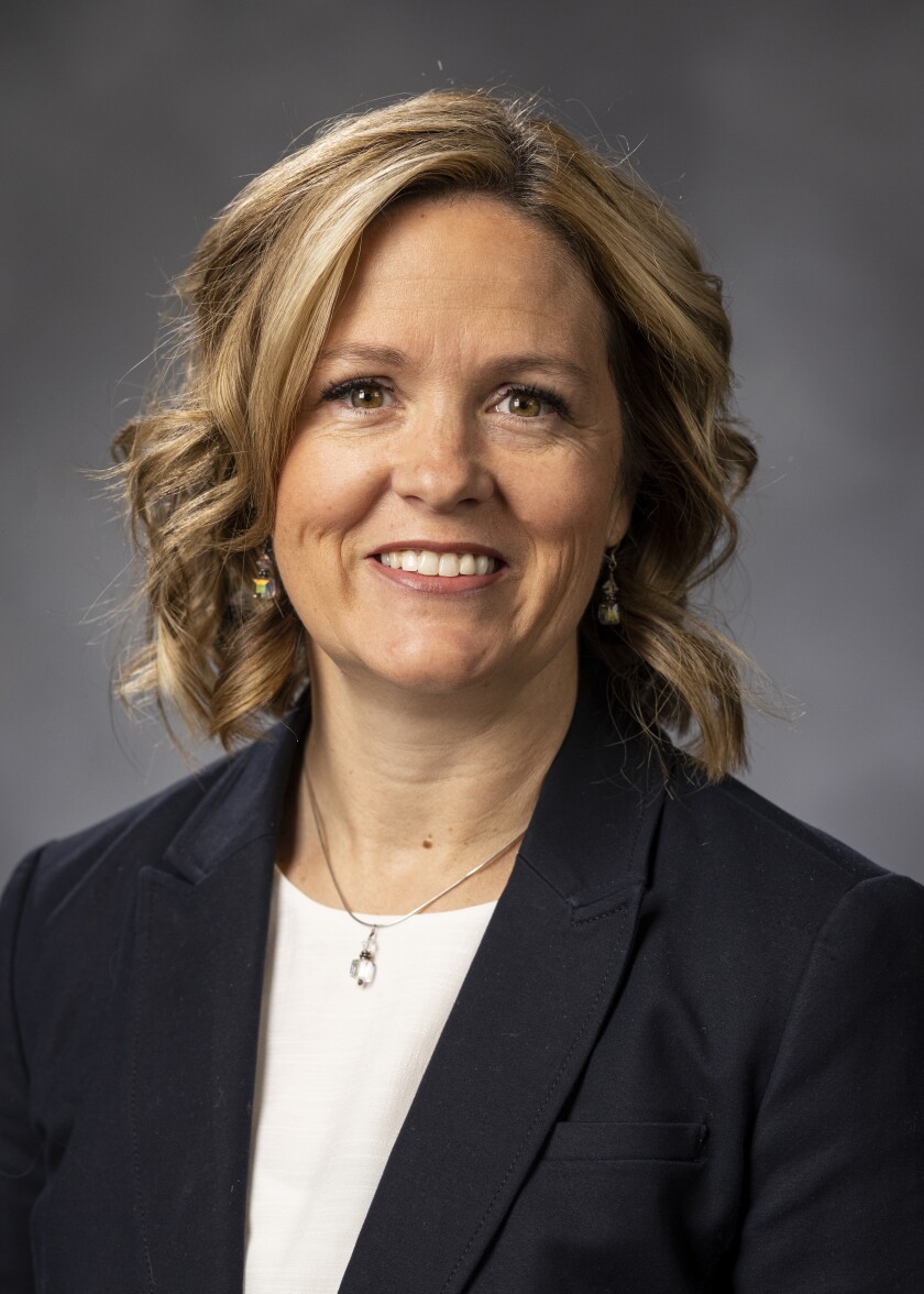 A woman with dark blonde hair wearing a dark suit coat and white shirt smiles for a professional headshot.