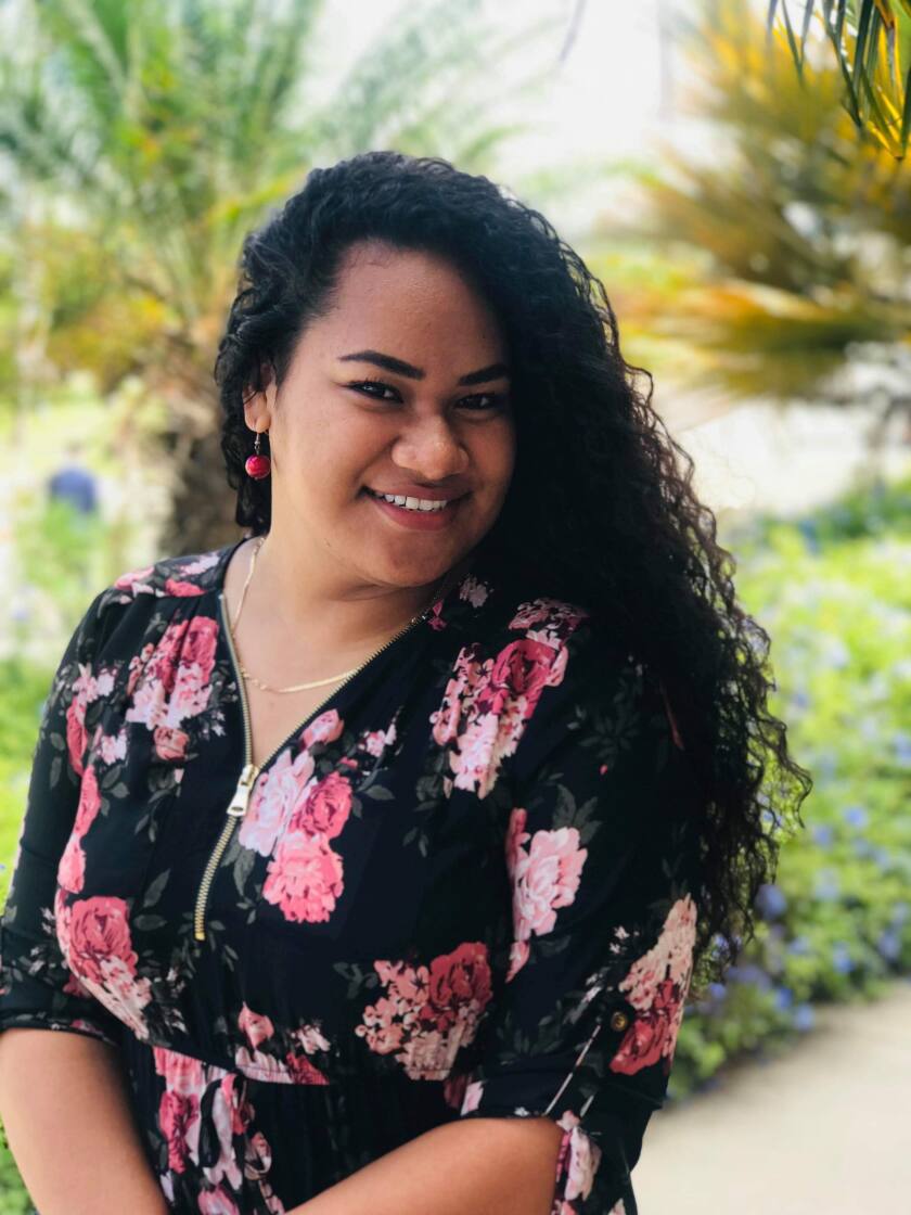 Amelia Otuafi standing with crossed arms in front of some greenery. She is wearing her hair down, a gold necklace, red earrings, and a black dress with pink flowers and a gold zipper in the front.