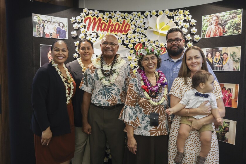 Irene Lesuma with six of her family members. They are all dressed in neutral formal attire and four of them are wearing white, green, purple and red leis. They are standing in front of a black backdrop with photos of Irene Lesuma, a sparkly red and yellow "Mahalo" and white and yellow flowers.