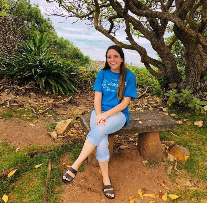 Diana Hawkins, wearing a blue shirt and pants, sits on a bench near the ocean.