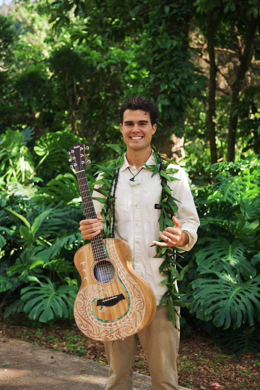A man stands outside holding a guitar in one hand and putting up a shaka in the other.