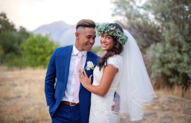 Malia wearing her wedding dress and Ojay Malala in a blue suit and pink tie with greenery and mountains in the background.