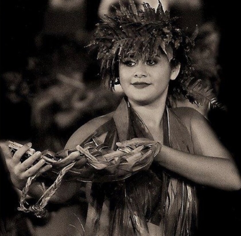 Tahitian woman dressed in traditional attire made from leaves, flowers and woven plant matter, holds up a woven basket in her hands while she dances.