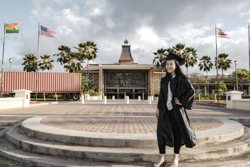 Muugii Galbadrakh smiling standing wearing a black graduation gown and cap with the BYUH campus and flags behind her.