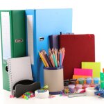 School and office supplies such as pencils, notebooks, folders, paint colors, tape, push pins, stapler, sticky notes, etc. on white background.