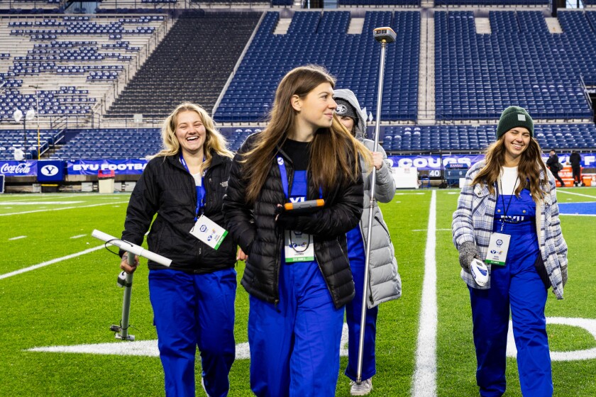 A group of students walk across a field with tools in their hands and blue matching overalls