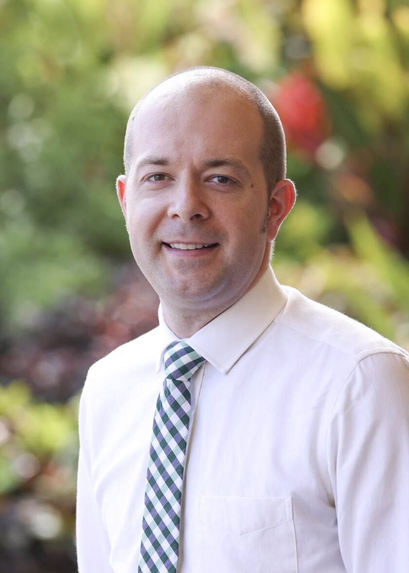 Jefferey Maloney, wearing a white long-sleeve dress shirt and blue and white plaid tie, stands in front of some blurred shrubbery.