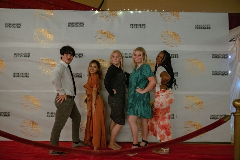 Five students pose in front of the same white, gold and black backdrop. The red carpet is also visible. The boy is wearing grey pants and shoes, a long sleeve white button-up shirt, and a black tie. One girl is wearing a long orange dress, another a short black and grey dress, another a short green dress, and the other a white shirt with a pink and red tie-dye skirt. They are all facing the side with their hands on their hips.