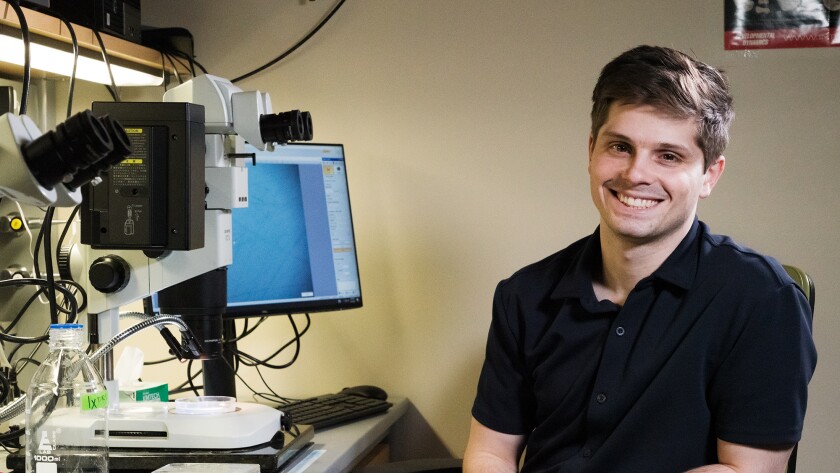 Jacob Wengler sits at a chair (right) smiling in a dark blue polo shirt, light skin, brown hair. To the left is a computer, microscope and other equipment.