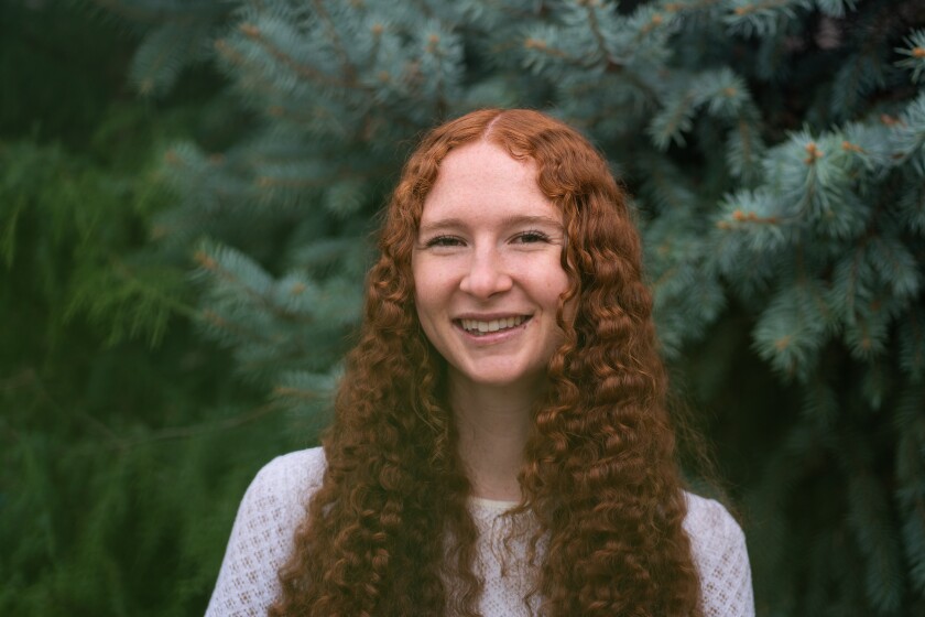 Maggie Briggs is a light skinned female with long wavy red hair. She's wearing a long sleeved white top and is posing for her headshot in front of some dark green fir trees.