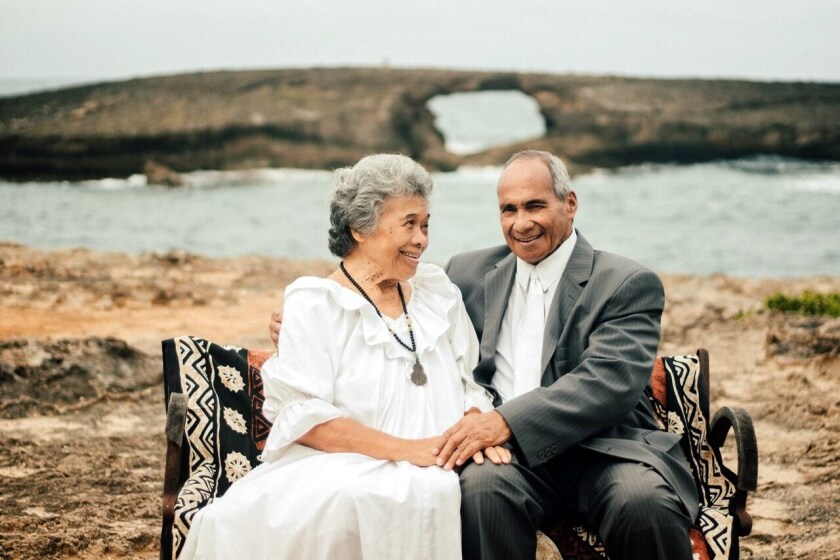 A older couple sits on  bench with the ocean and rocky shoreline behind them. The woman is wearing  white muumuu and the man a grey suit, and a white shirt and tie.