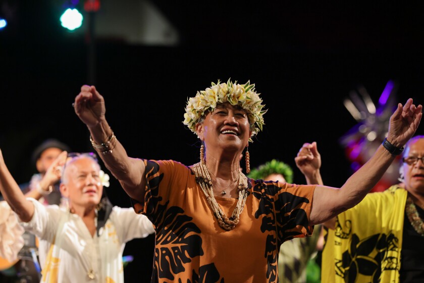 Kekela Miller is doing the hula wearing an orange dress with leaves, beaded jewelry and a flower haku on her head with other women behind her doing it too.