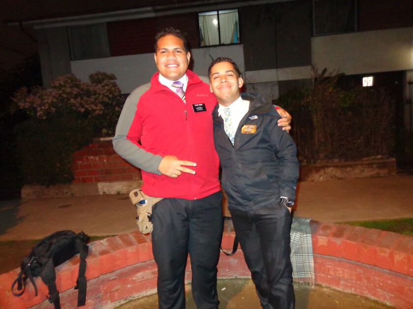 Lionel Funaki, wearing black pants, pink tie, and a gray and red pullover with his missionary badge. His companion is wearing a black jacket, black pants, and a floral tie. They are standing near a sidewalk with their arms around each other.
