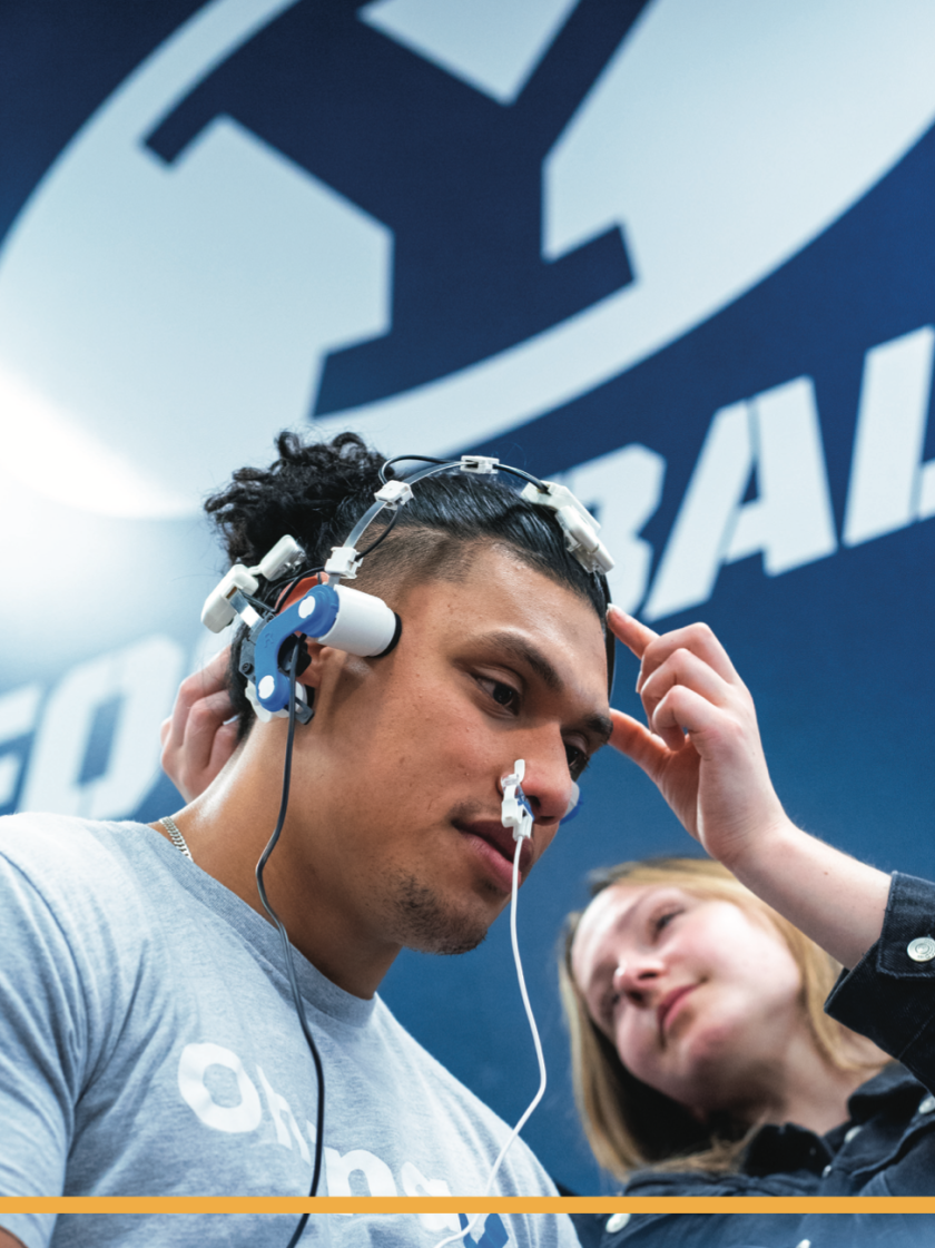 A white female student adjusts wires on a football player's head.