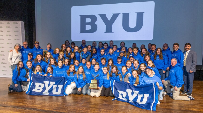 Four rows of smiling male and female college students wearing blue hoodies posing together for a photo. The students in the first two rows are kneeling on the floor of the stage. The student in the center of the first row holds a trophy while the students on the ends of the row hold BYU flags. Projected onto the screen behind them are the letters 'BYU'.
