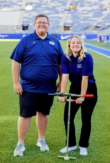 An older man stands to the left. A young, blonde woman stands to the right. She is holding a silver tool.