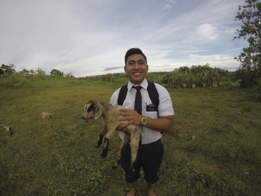 A missionary wearing a white dress shirt, dark tie and pants, wears a backpack and holds a goat while standing in a grassy field in the Philippines with banana plants in the background.