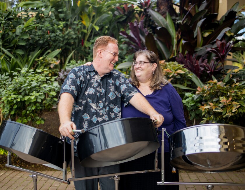 The Duerdens stand looking at each other, Darren wearing a aloha shirt with blue fish and Jennifer with a dark blue shirt with shaka steel drums in front of them.