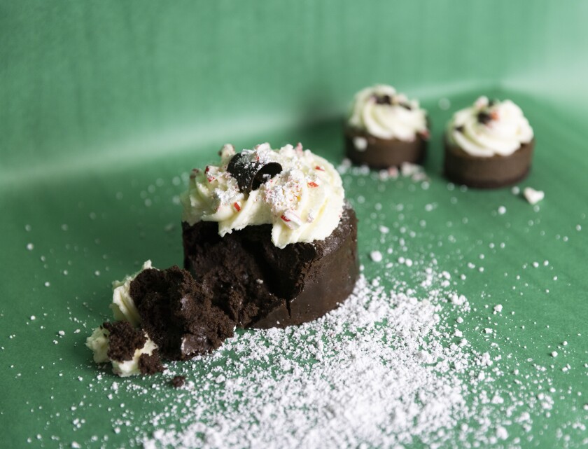 A small chocolate cake with white icing, powdered sugar, candy cane pieces and chocolate on top. One bite is taken out and sitting next to the cake, which is placed on a green background with powder sugar around it.
