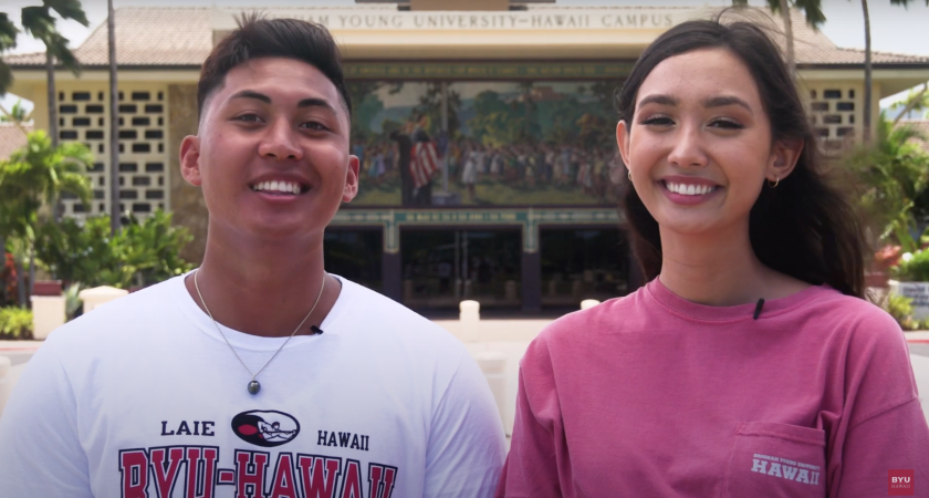 A man wearing a white BYUH shirt stands beside a woman in a pink BYUH shirt with the BYUH campus flag raising mural in the background.