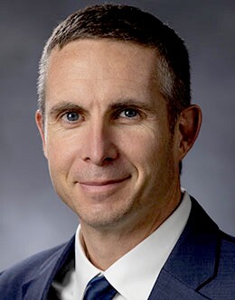 A pale man with greyish hair smiles while wearing a suit, white shirt, and tie.