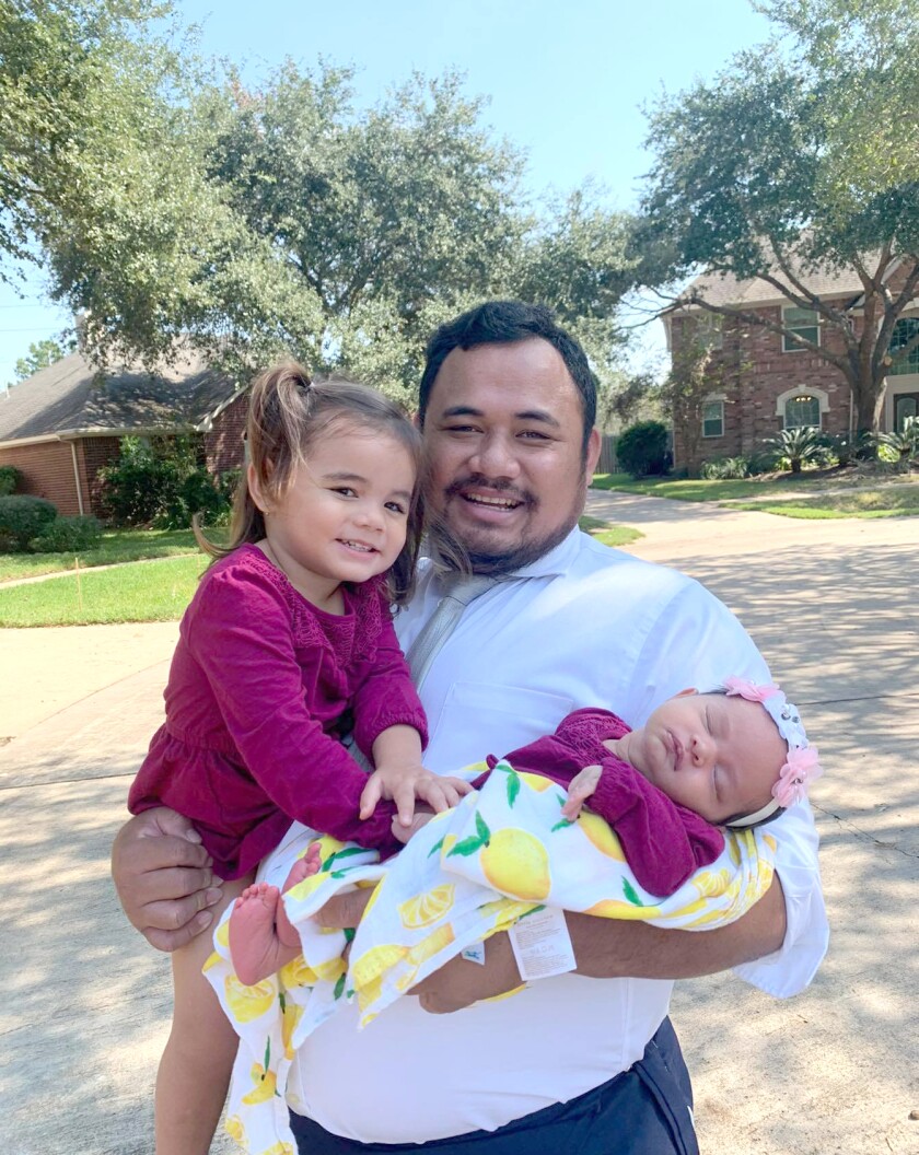 Siona is smiling and holding a sleeping baby girl wearing red wrapped in a white blanket with lemons in his left arm and another toddler girl wearing red in his right arm.