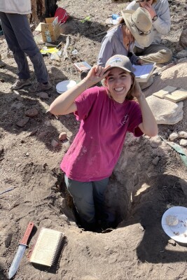 A woman adjusts her tan baseball cap with both hands with a wide smile, one hand on the brim and one of the back of her head. She is standing hip-deep in a hole, wearing gray work pants and a fuschia tshirt. Around her on the ground are a spiral notebook, a knife, and a paper plate with various rocks. Behind her two people stand out of frame and another two are cross-legged on the ground reviewing their own notebooks.