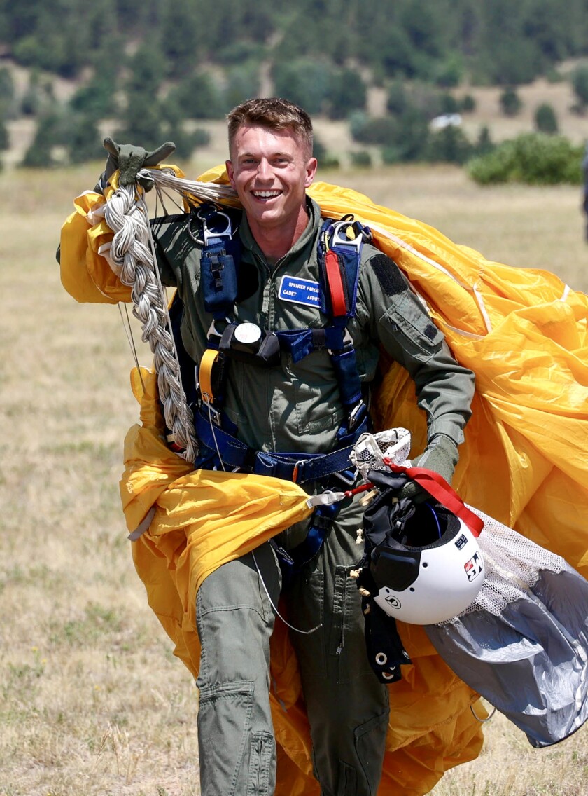 Parker smiles while walking with a yellow parachute over his shoulders. He wears an air force jumpsuit and carries his gear.