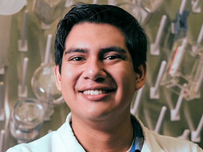 A hispanic man in a lab coat smiles in front of lab test vials