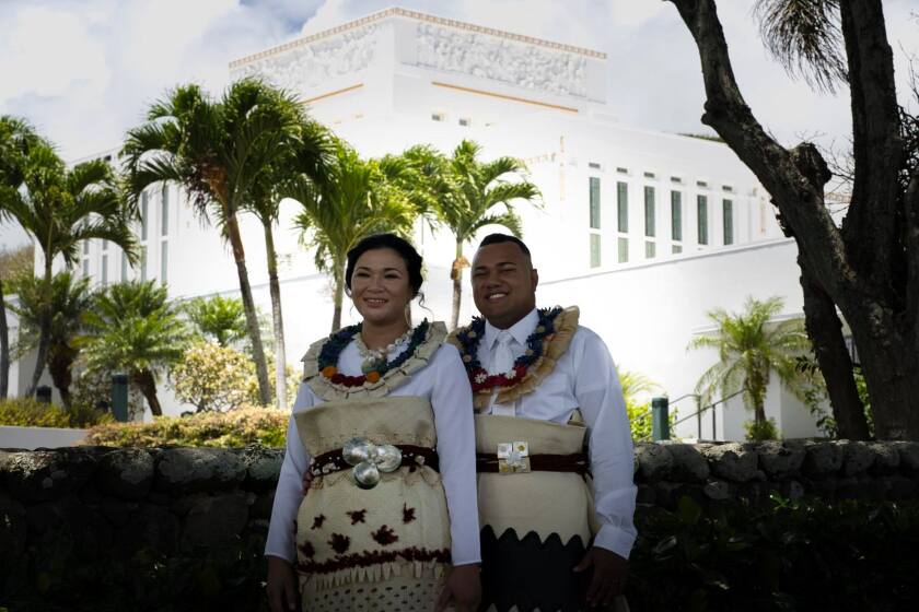 The Senilolis stand together in white temple clothes in front of the Laie Hawaii Temple wearing traditional Tongan leis and mats with mother of pearl and other embroidery.