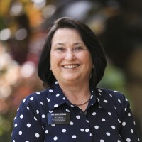 A portrait of Sister Larson smiling at the camera. Plants are blurred in the background.
