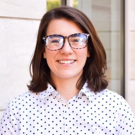 Emma Belnap smiles for a headshot. She is standing outside and wearing glasses and a polka dot button-up blouse.