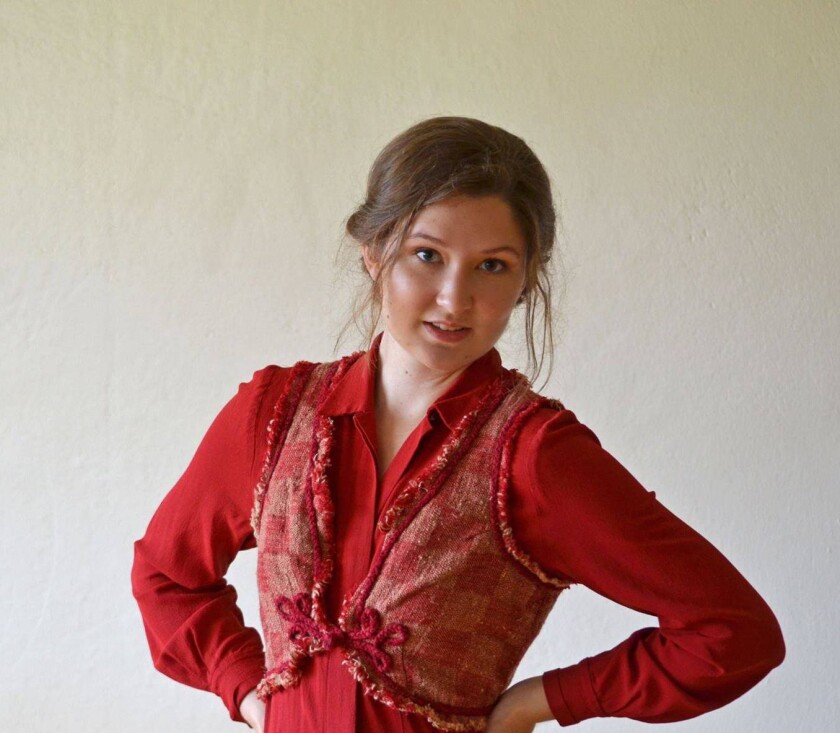 Kiaran Loefke wears a red long-sleeved blouse with her hair tied up behind her with a white-walled background.