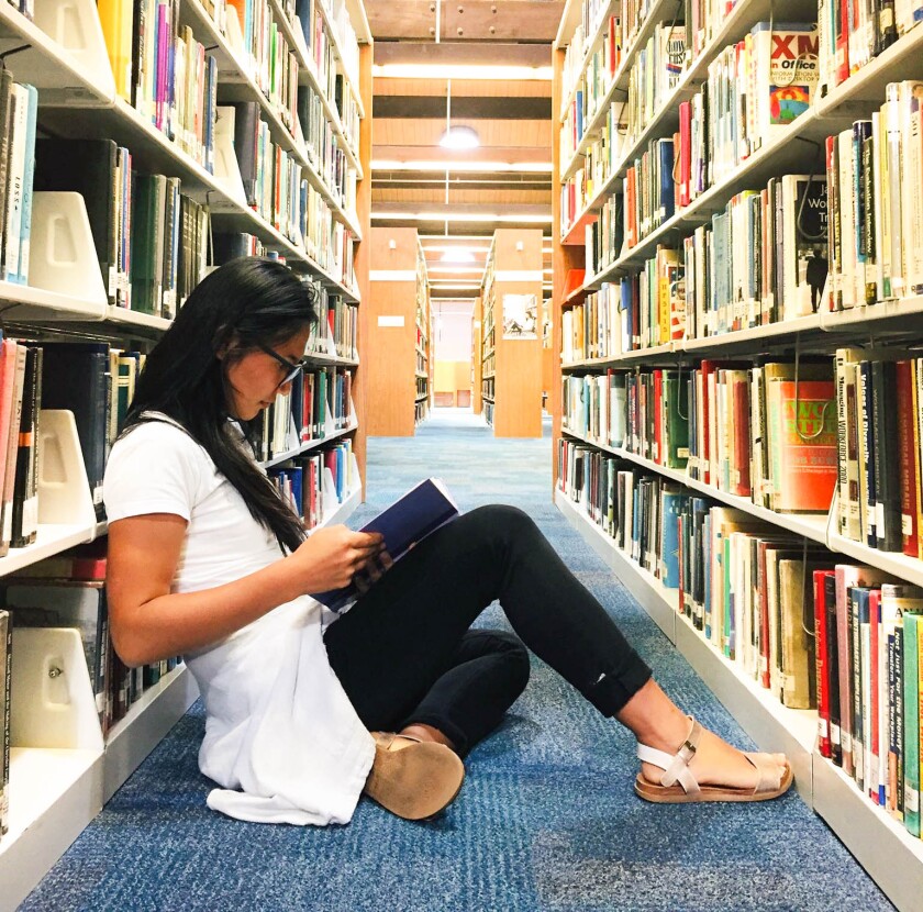 Dalaichuluun sitting on the ground in the library between two bookshelves reading a book.