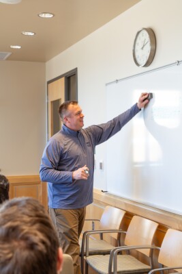 A male professor in a blue BYU pullover stands at a whiteboard with an eraser.