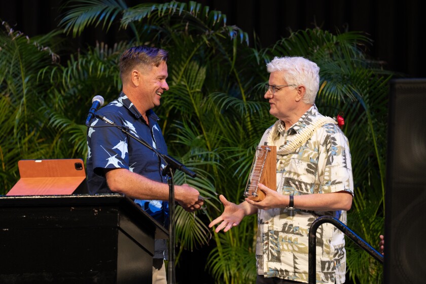 One man holding a plaque and wearing a lei shakes hands with another man at a podium.