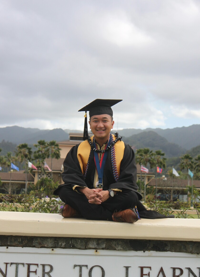 Young man in graduation gown and cap sits in front of BYU-Hawaii.