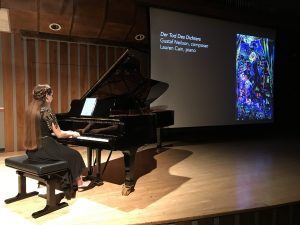 A young woman in a black dress plays the piano with an image of abstract art behind her.
