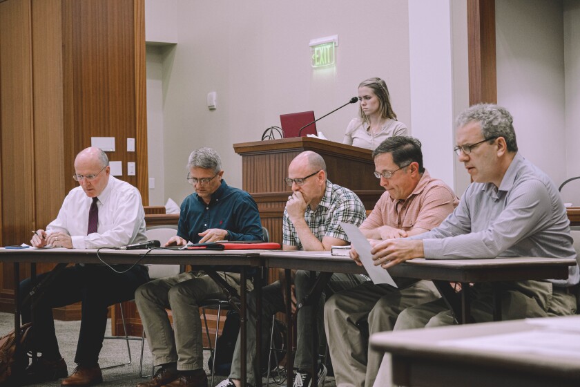 Five professors sitting by the table and a lady standing behind the podium.