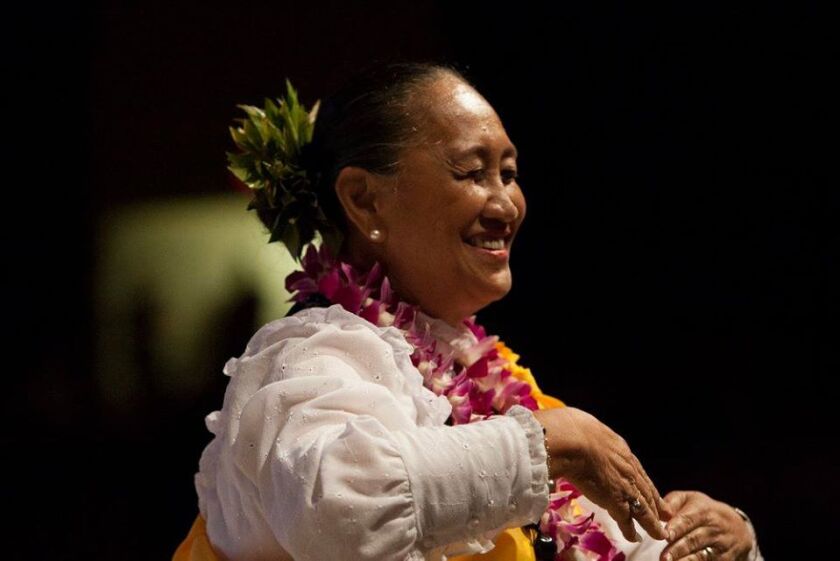 Aunty Kela smiles as she's performing the hula in a white shirt with leis and black background.