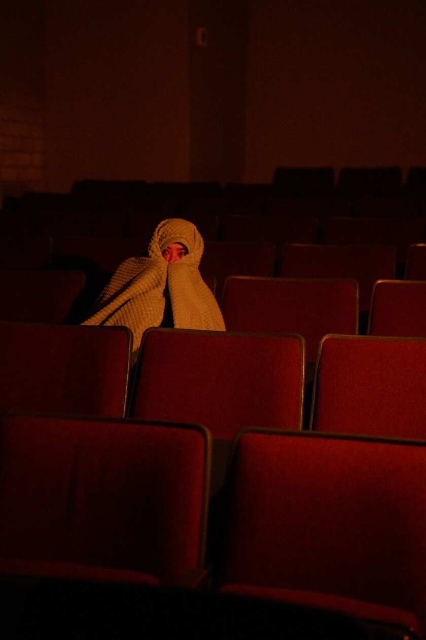 A woman sits with a blanket over her head and her eyes peeking out in the red seats of a darkened movie theater.