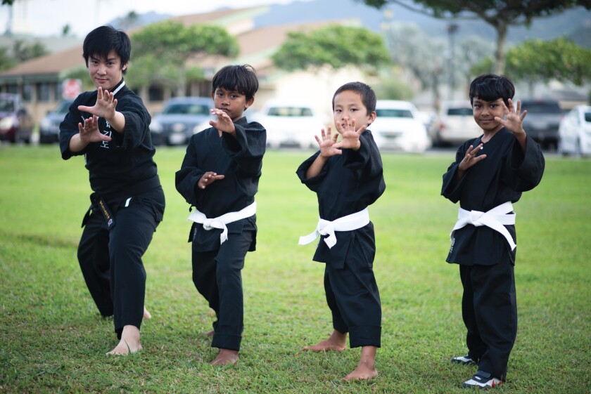 A Hopkido teacher and three young boy students all wearing black tops and pants, strike a martial arts pose on the grass outside of the apartments they live in on campus.