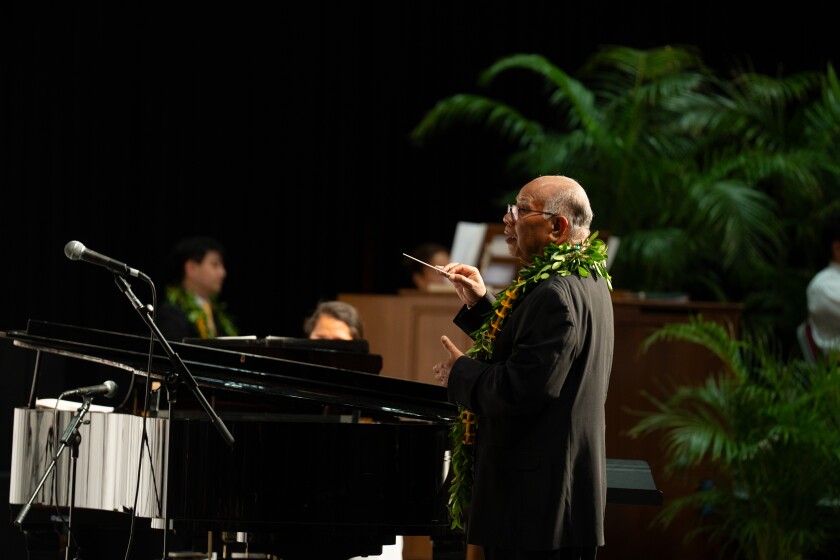A man in a black suit coat and wearing a leafy green lei stands behind a piano with his arms up conducting a choir.