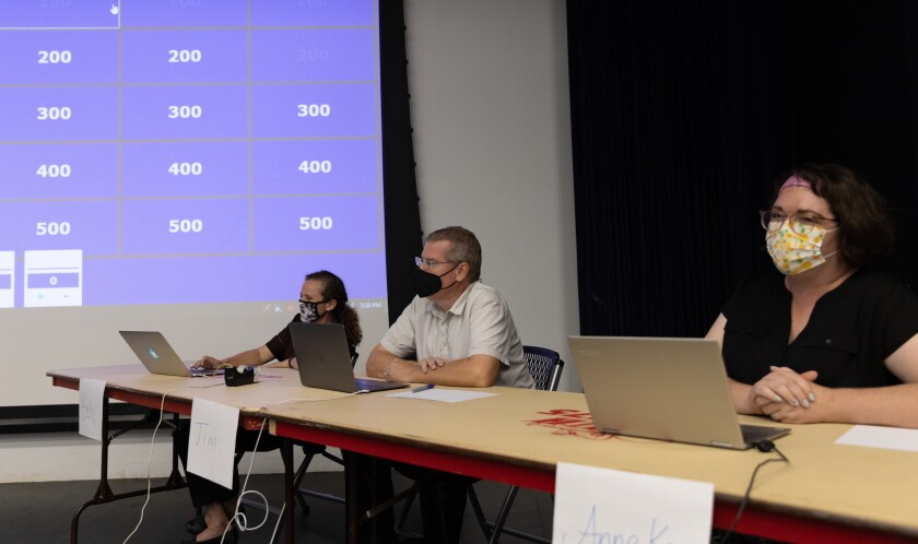 Becky Strain, in a black shirt, Jim Tueller, in a dark white button-up, and Anneke Garcia, wearing a black shirt, sit at the jeopardy table with the jeopardy game in the background on a big screen.