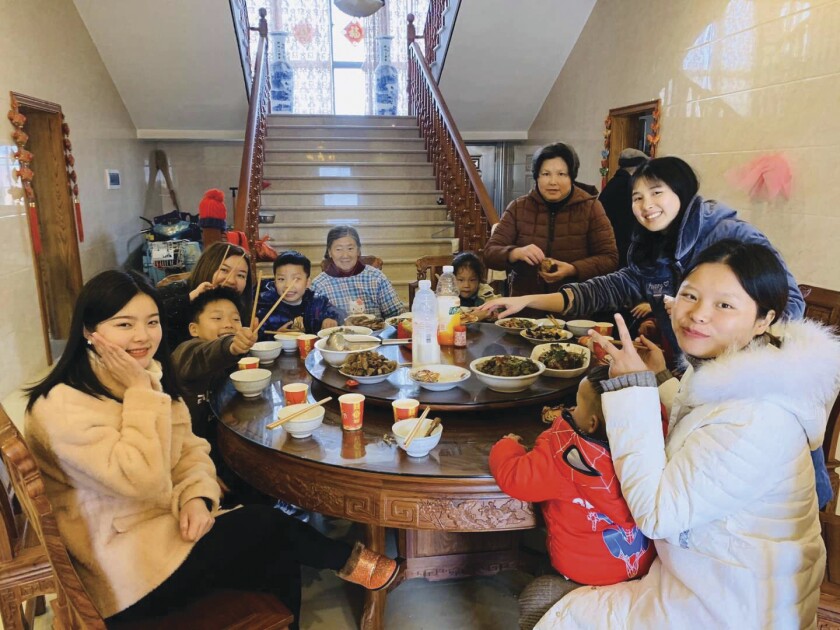 Fei Liu and her family of six women and four children sit around a large, round table eating a meal in white bowls of rice and a variety of main dishes.