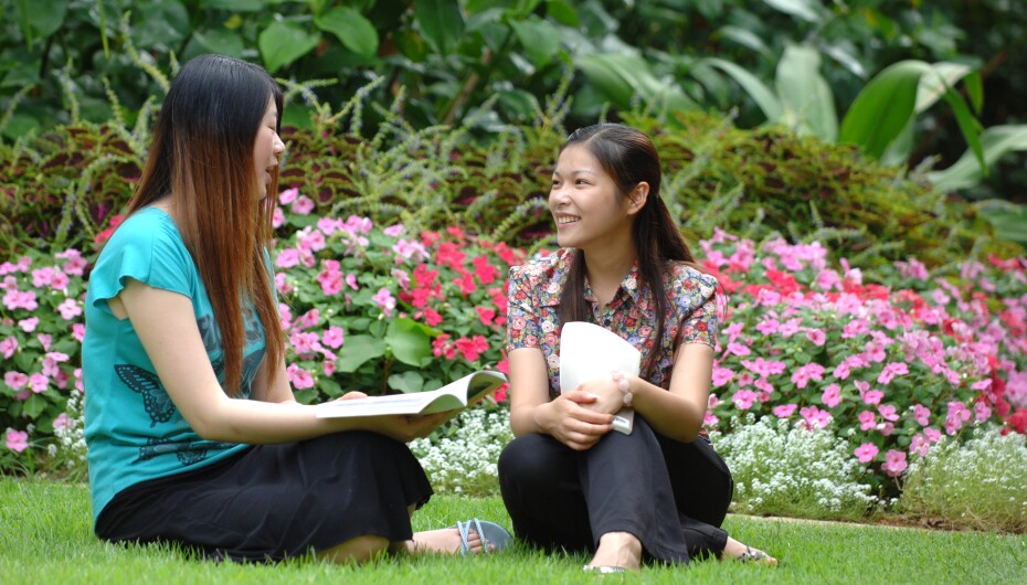 A picture of two female students sitting in a green grassy lawn talking.