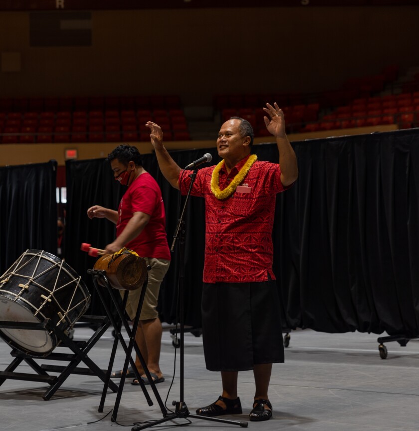 Man in red shirt and yellow lei stands at a microphone on the floor of the CAC with his arms up and a man with drums standing behind him