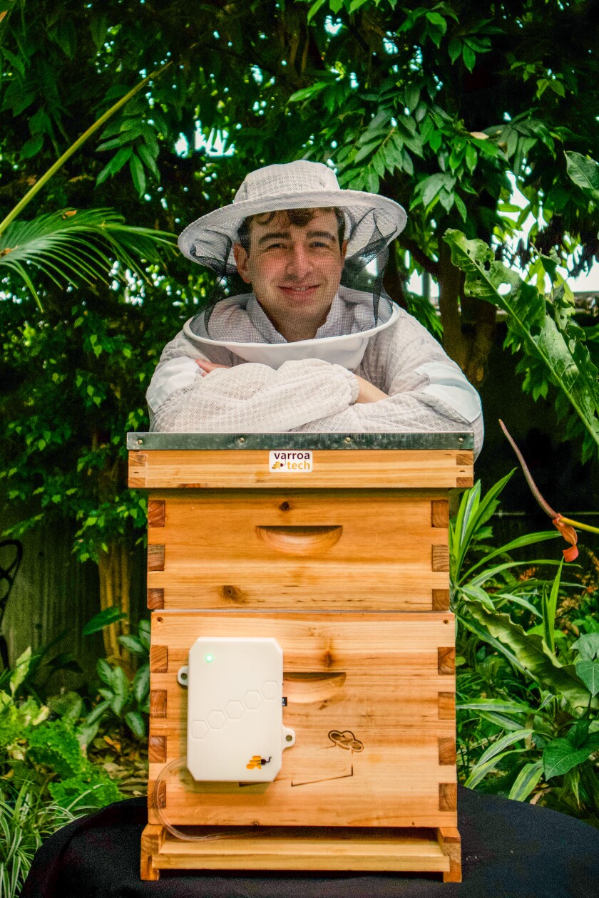 A male college student wearing a beekeeping suit poses with the new product. The product is a light brown wooden box with a small white device fastened on the bottom left corner.