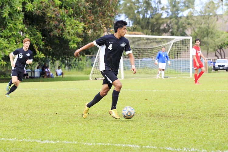 A male soccer player wearing a black uniform and yellow cleats kicking a soccer ball across a field